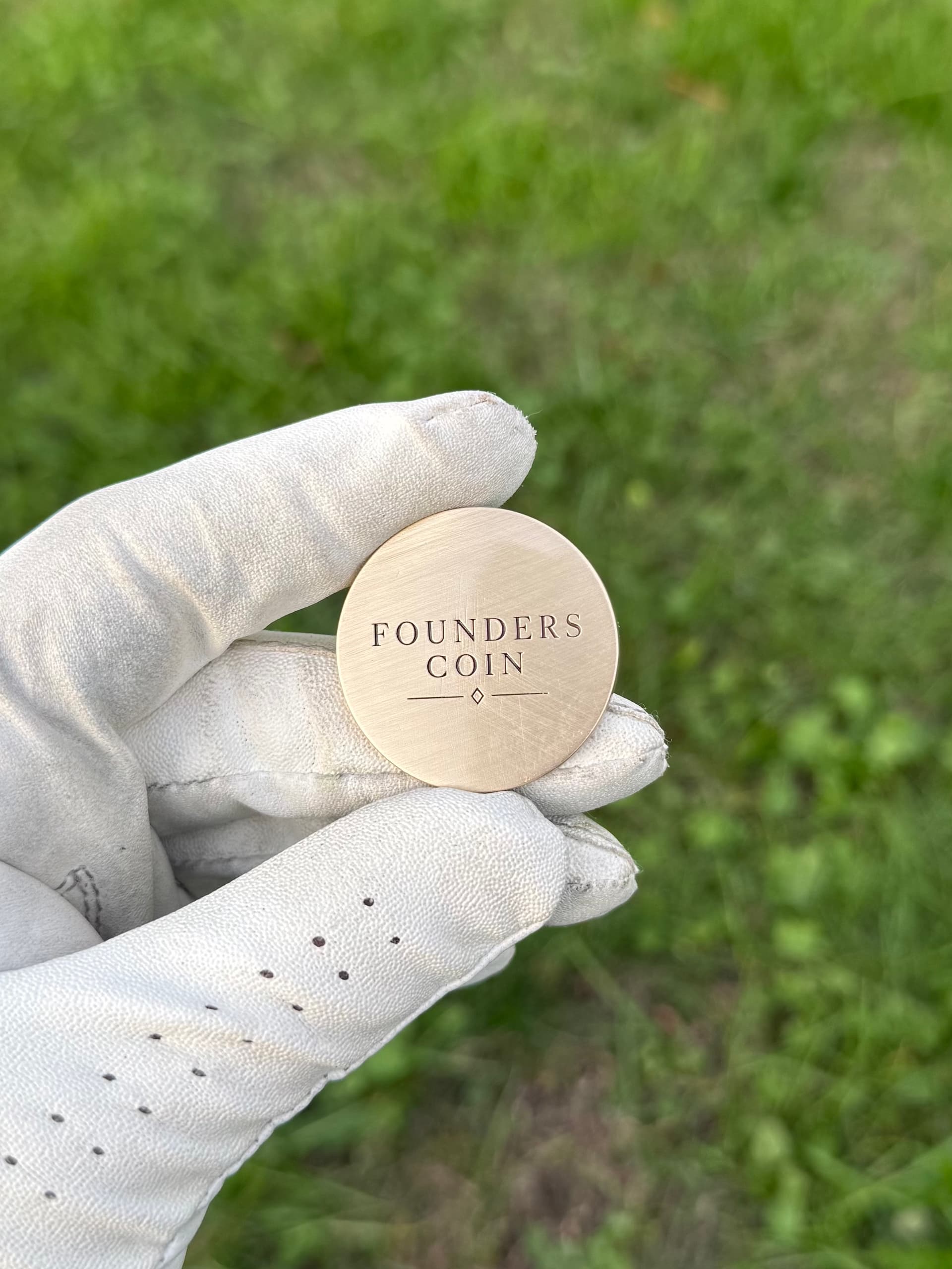 Gloved hand holding a solid brass Founders Coin golf ball marker