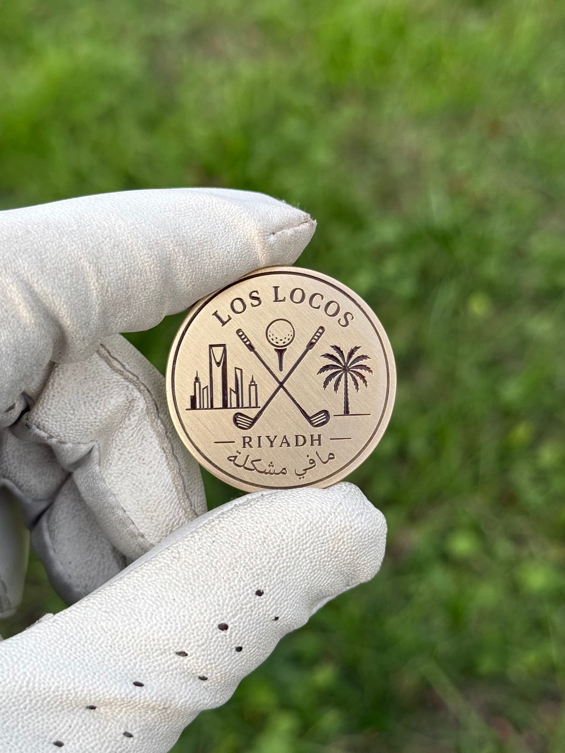 Gloved hand holding a solid brass golf ball marker engraved with the Los Locos Riyadh crest